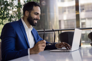 Handsome smiling arabic businessman planning start up project, typing on keyboard in modern office. Confident middle eastern man wearing suit holding eyeglasses working online, sitting at workplace - obrazy, fototapety, plakaty