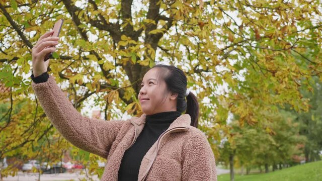 Happy Asian woman taking a selfie on smartphone at the park in Autumn, woman wearing light brown coat, yellow leaf on the tree, Beautiful day in Autumn season, Sweden