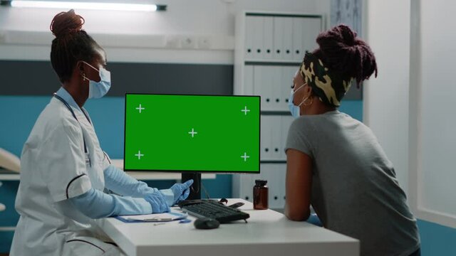 African american doctor and patient looking at green screen on computer in cabinet. Black medic and woman watching monitor with mockup template and isolated background for chroma key.