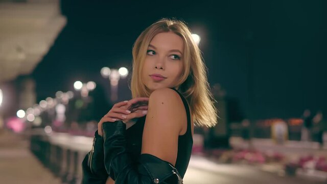 Portrait of a charming blonde in the light of the street lamps of the night city. Sexy girl posing and flirting with the camera