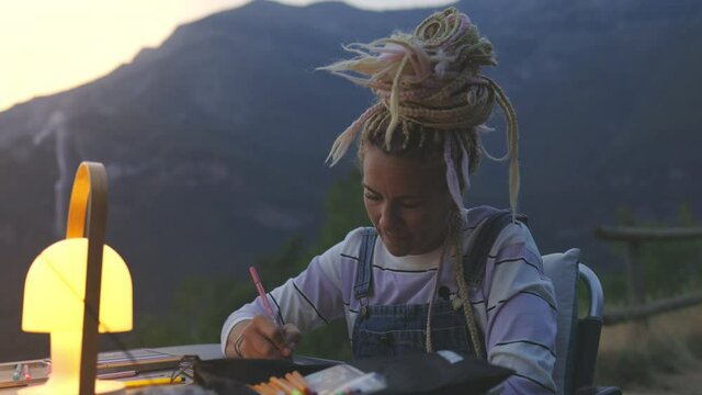 Hippy girl with blond dreadlocks, deep in thought draws or writes outdoors