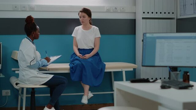 African american doctor talking to patient on bed at annual checkup visit. Black medic and caucasian woman doing medical consultation in doctors office for healthcare. Adult at examination
