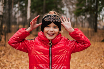 Laughing little girl in autumn forest making faces sticking out her tongue. Happy child goofing, playing. Smiling kid having fun outdoors. Red jacket, knitted headband, black pants.