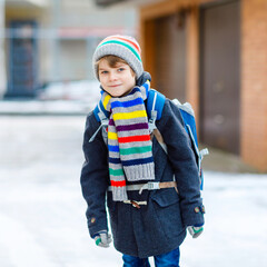 Little school kid boy of elementary class walking to school during snowfall. Happy child having fun and playing with first snow. Student with backpack or satchel in colorful winter clothes.