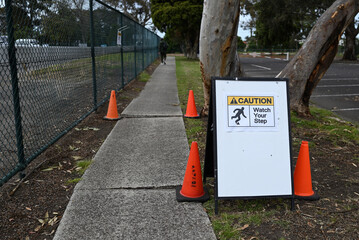 A caution watch your step sign, warning about uneven pavement on a footpath, with orange traffic cones. An out of focus person can be seen walking the path in the background