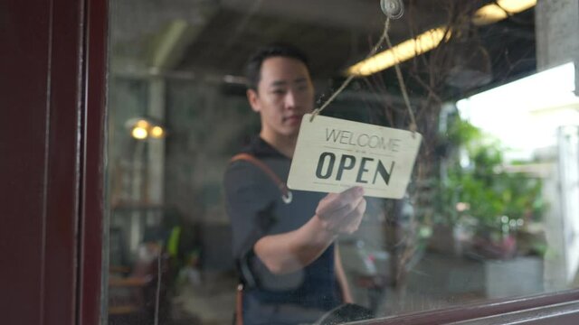 Asian man coffee shop barista walking to cafe door and turning hanging closed sign to open. Male waiter preparing restaurant service to customer. Small business owner and part time job working concept