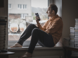 A man with a mobile phone sits on a window sill surrounded by books.