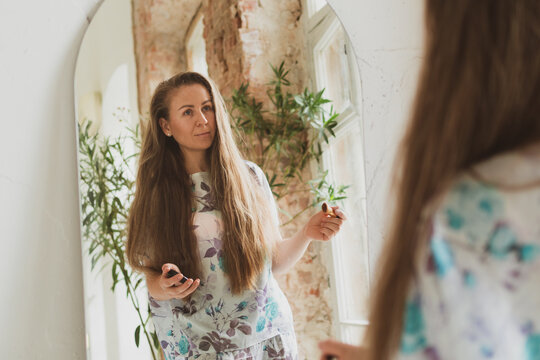 young woman with long hair combing her hair and applying makeup near the mirror