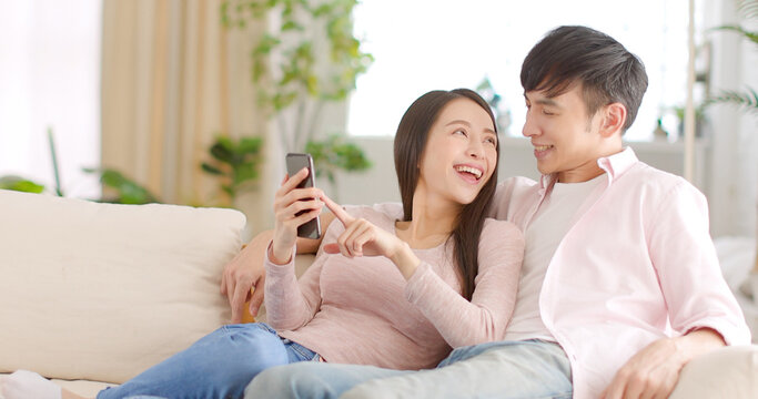 Happy Young Couple Sitting On  Couch And  Looking At Mobile Phone