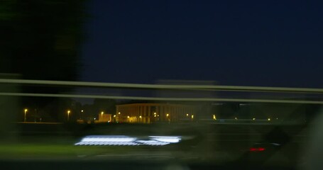 Car quickly passes along road of night town, past avenues and buildings illuminated by street lamps, fenced city parks, view through window from inside automobile. Other vehicles are rushing past.
