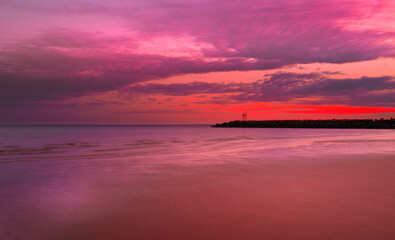 Obraz premium Dramatic pink and red seascape at dawn with dark clouds illuminated by the rising sun. Soft misty sea waves rolling in on the sandy beach reflecting warm glowing light. 