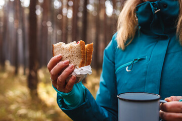 Woman get hungry during hiking in forest. Female tourist eating sandwich and drinking hot tea...
