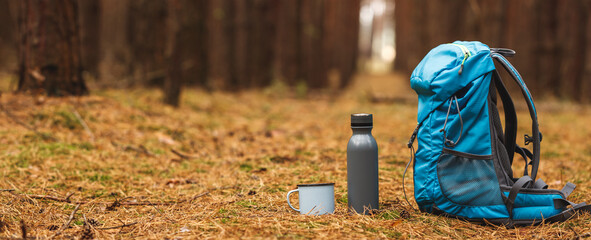 Backpack, thermos and travel mug in forest. Hiking equipment on footpath in woodland. Panoramic view