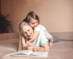 Home comfort. Grandma reads book to granddaughter on sofa. Child gently hugs grandmother. Connection generations. Beige interior.Family holiday and togetherness.Spend time together