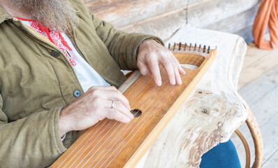A man in ethnic clothes plays an ancient musical instrument. A bearded man, a forester has built a house and is playing a psaltery.