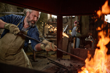 Professional smith in gloves burning the flame in the furnace with adding coals