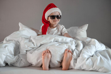 Funny cute boy is sitting on the bed in sunglasses, sipping in a Santa hat. Happy holidays....