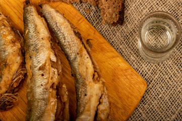 A small fried fish on a wooden board, pieces of bread and a glass of vodka, close-up, selective focus.