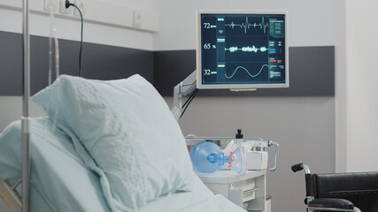 Close up of heart rate monitor in empty hospital ward. Nobody in intensive care room with medical equipment, bed, oxygen tube and wheelchair for recovery. Healthcare instruments
