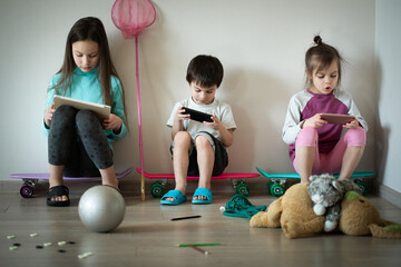 Three funny serious children sitting on skateboards and playing mobile games on gadgets at home