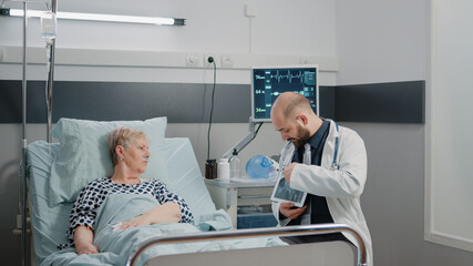 Doctor holding tablet with x ray scan for healthcare examination. Old patient with disease looking at radiography on gadget screen for diagnosis and treatment in hospital ward bed.