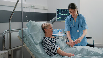 Medical assistant putting oximeter on finger of aged patient in hospital ward bed for oxygen saturation measurement and pulse pressure. Nurse helping retired woman with IV drip bag