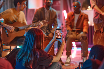 Rear view of young woman shooting the performance of musical band on her mobile phone in the club