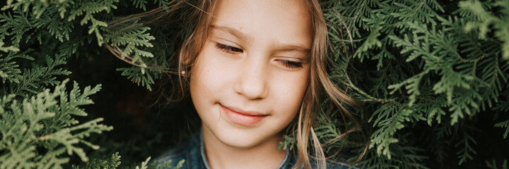 portrait of the face of a cute happy caucasian candid healthy eight year old kid girl surrounded by branches and leaves of green plant thuja or cypress on nature outdoor. banner