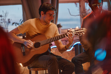 Young man sitting on chair and playing guitar for his friends in the room