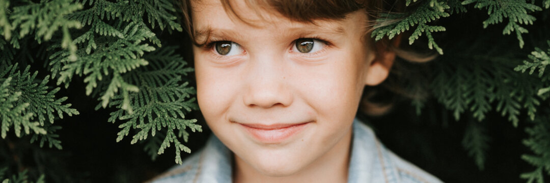 portrait of the face of a cute little happy caucasian candid healthy five year old kid boy surrounded by branches and leaves of green plant thuja or cypress on nature outdoor. banner