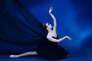 Portrait of adorable female ballet dancer in black tutu and stage make-up sitting on floor isolated on blue studio backgorund