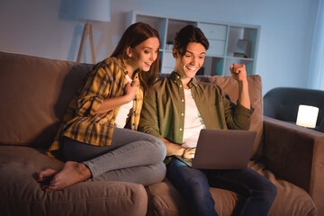 Photo of pretty lucky lovers dressed casual shirts sitting sofa hugging looking modern device rising fists smiling indoors room home