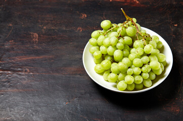 Branch of ripe green grape on plate with water drops. Juicy grapes on wooden background, closeup. Grapes on dark kitchen table with copy space