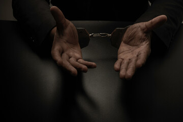 A businessman in a black suit, handcuffed on a table and a black table, is making gestures by...