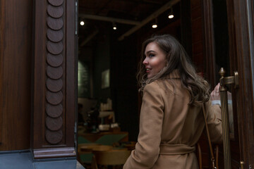 Obraz na płótnie Canvas A young woman walks through the door to a restaurant. Smiling beautiful blonde in a beige coat.