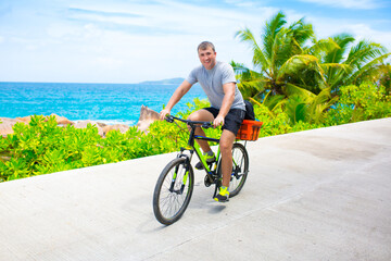 Fototapeta premium Happy young guy riding a bike on the road by the sea in the Seychelles. Concept of vacation and active lifestyle. Cycling. The man on the bike.