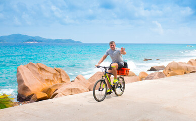 Fototapeta premium Happy young guy riding a bike on the road by the sea in the Seychelles. Concept of vacation and active lifestyle. Cycling. The man on the bike.