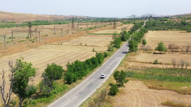 Aerial view car on highway, between fields.  Aerial footage white, soviet style Russian car along the rural highway on road. Zhiguli, Lada, old style car on traffic in Armenia. Near cultivated lands.