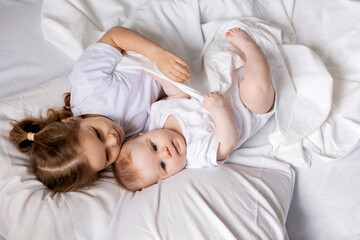 Obraz na płótnie Canvas little girl and baby in white clothes are lying in bed on white bed linen. brother and sister bask in bed. children's sleep and rest. hygge, lifestyle. space for text. High quality photo