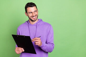 Photo of optimistic millennial brunet guy write paper look wear spectacles hoodie isolated on green color background