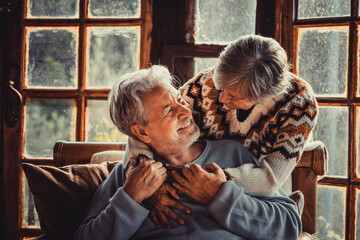 Happy senior couple enjoy with love winter holidays at home hugging with joy and romance. Romantic elderly lifestyle. Man and woman embrace and look each other at home with snow outside