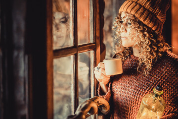 Adult thoughtful woman look outside the window drinking coffee at home. Female people reflected on the glass looking outside waiting and thinking. Winter holiday season lifestyle concept