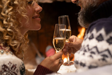 Close up of happy couple in love clinking flutes of wine with fired fireplace in background. Romantic people enjoying relationship at home during winter holidays. Concept of man and woman together