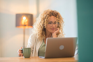 Beautiful caucasian young woman with curly hair and eyeglasses drinking coffee and looking at...