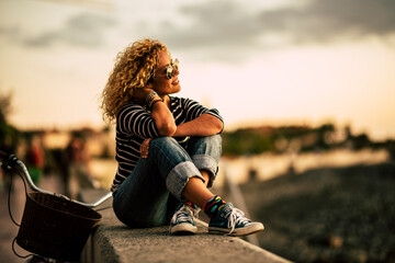 Hipster woman with curly hair and sunglasses sitting on embankment wall and admiring beautiful...