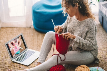 Young woman knitting wool using needle while watching online tutorial on laptop at home. Woman watching needlework lessons, a laptop and home hobbies. Caucasian woman learning to knit online - obrazy, fototapety, plakaty