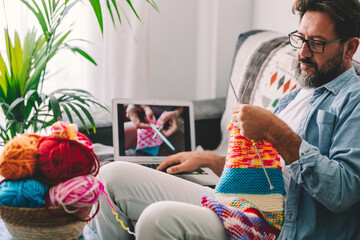 Mature man knitting wool using needle while watching online tutorial on laptop at home. Man looking at knitted wool while relaxing of sofa. Caucasian man learning to knit online
