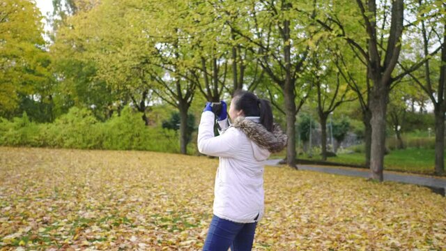 Asian woman taking a photo of view at park. Walking around with Autumn outfits, Good weather, yellow leaves falling on the grass. A lot of tree background, Stockholm, Sweden in Autumn concept