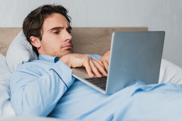 Young freelancer in pajama using blurred laptop while lying on bed in morning