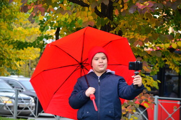 a schoolboy teenager in a warm jacket and a hood walks in an autumn park with colorful foliage with red umbrella. records video to phone. modern youth. blogger. communicates online, streams
